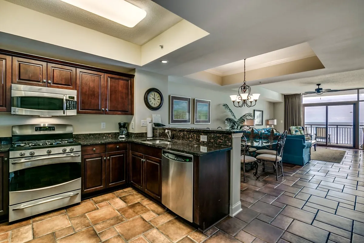 An open kitchen and dining area with sleek cabinetry and stainless steel appliances. The dining table is placed to enjoy views through sliding doors, which open out to a balcony with glimpses of the ocean beyond. The space is airy and designed for shared meals and entertaining.