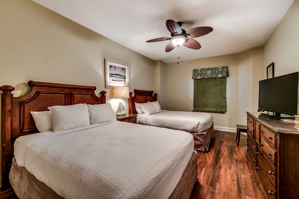 A cozy guest bedroom with two comfortable beds, a ceiling fan, a dresser, a TV, and a window adorned with green shades. The room feels welcoming and is well-suited for guests seeking comfort and entertainment.
