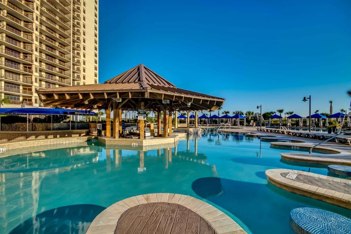 A vibrant outdoor pool area featuring a gazebo, plentiful lounge chairs, and a lively atmosphere. The setup suggests spaces for social gatherings, sunbathing, or enjoying drinks by the pool, with hints of a swim-up bar and evening entertainment options.
