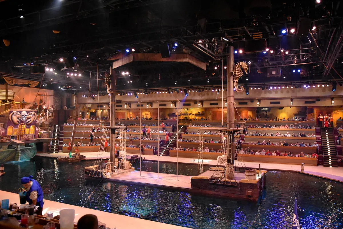 A vibrant interior shot of a live performance venue featuring a dramatic stage set over water, rows of audience seating, and theatrical props. The scene is lively, hinting at the excitement of immersive shows and dinner experiences.
