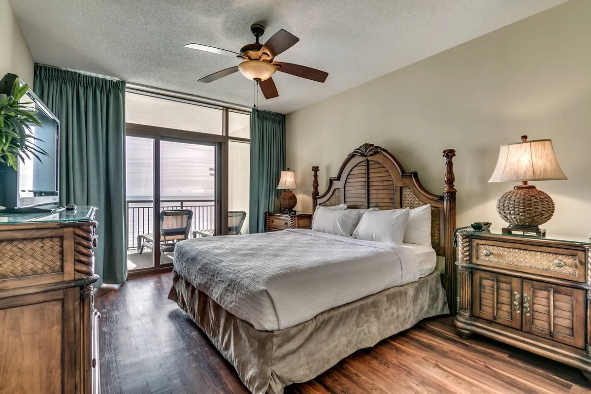 A spacious bedroom featuring a plush king bed with a decorative headboard, wooden furniture, and a large window that opens onto a balcony. Natural light floods the room, creating a tranquil and airy atmosphere ideal for restful moments.