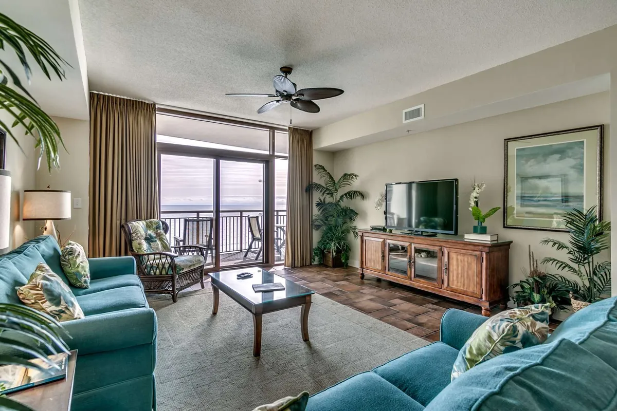 A sunlit living room featuring plush teal sofas arranged for conversation, a wooden TV cabinet, tropical plants for a beachy touch, and expansive windows offering sweeping ocean views. The scene exudes a tranquil, cozy atmosphere perfect for relaxing with friends or family while watching the waves or a sunset.