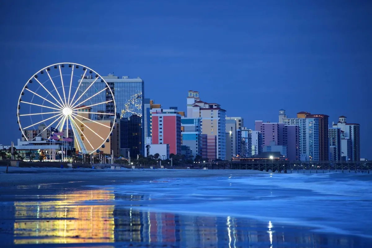 A lively beachfront scene featuring a Ferris wheel, vibrant buildings, and the reflective surface of the water. Visitors can be seen strolling along the beach, with amusement park rides and nearby restaurants adding to the bustling atmosphere.