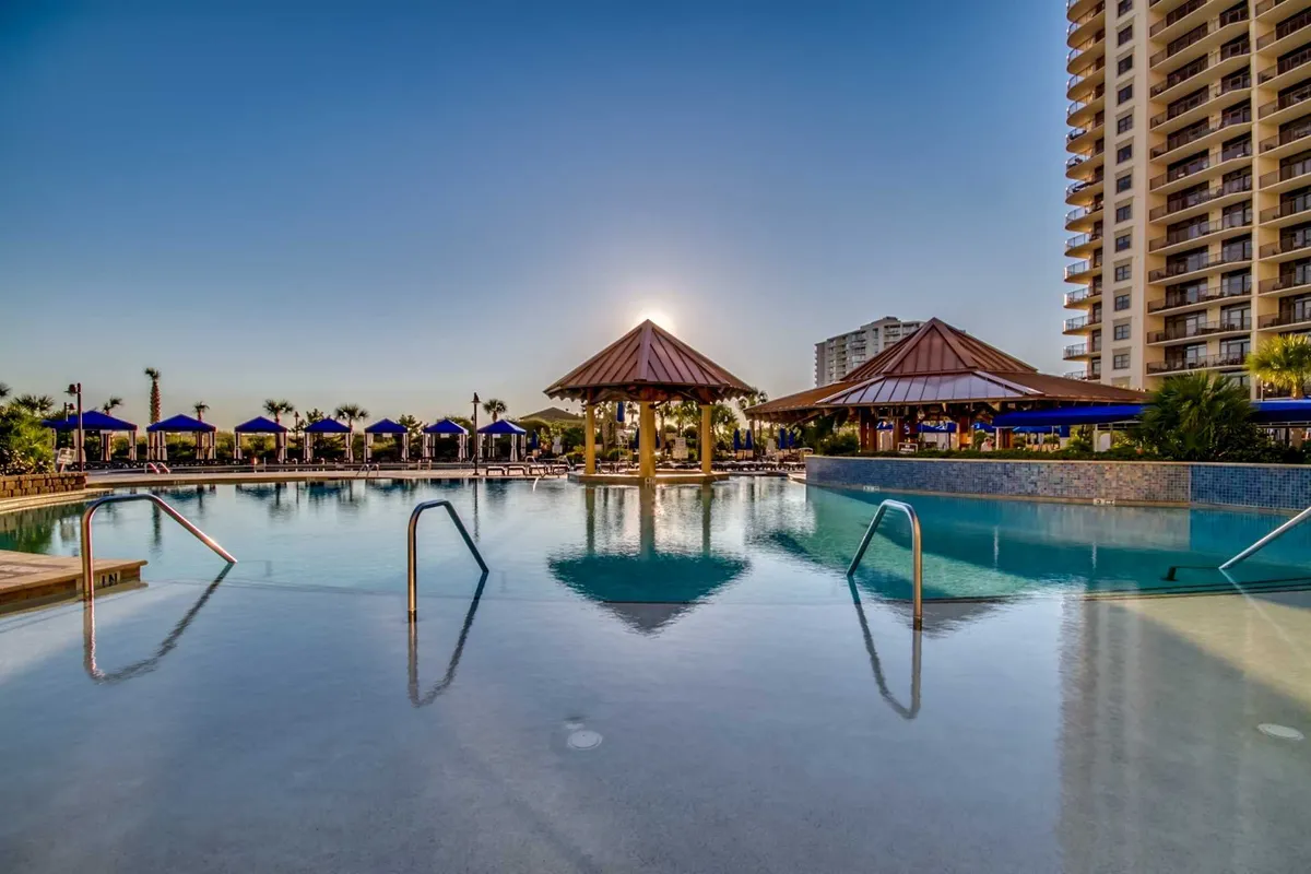 A tranquil scene at dusk with the resort pool in the foreground, framed by thatched-roof cabanas and lounge chairs. The setting sun casts a serene glow over the water, creating an inviting ambiance for relaxation, socializing, or enjoying a cocktail by the pool.