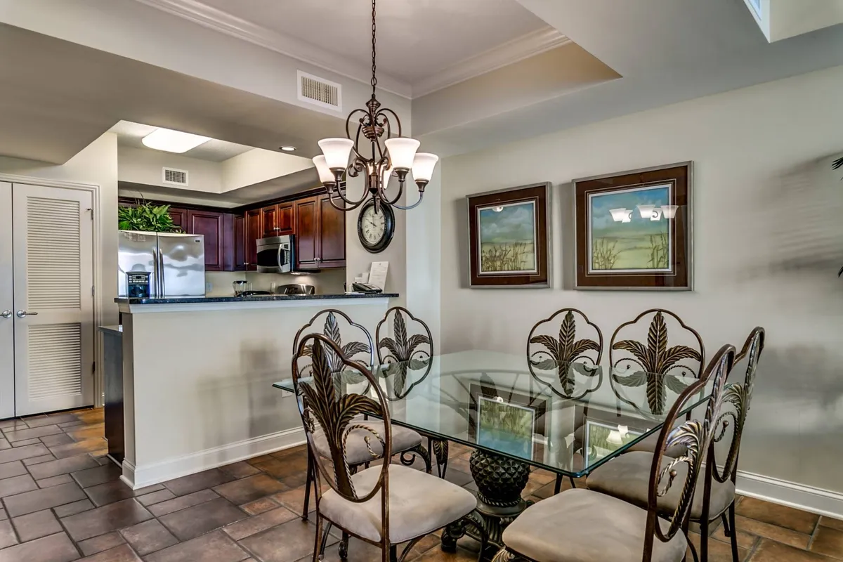 A sophisticated yet cozy dining nook with a glass dining table, ornate chairs, chandelier, and art on the walls. The open layout provides a view into the adjacent kitchen area, fostering a sense of connection between cooking and dining spaces.