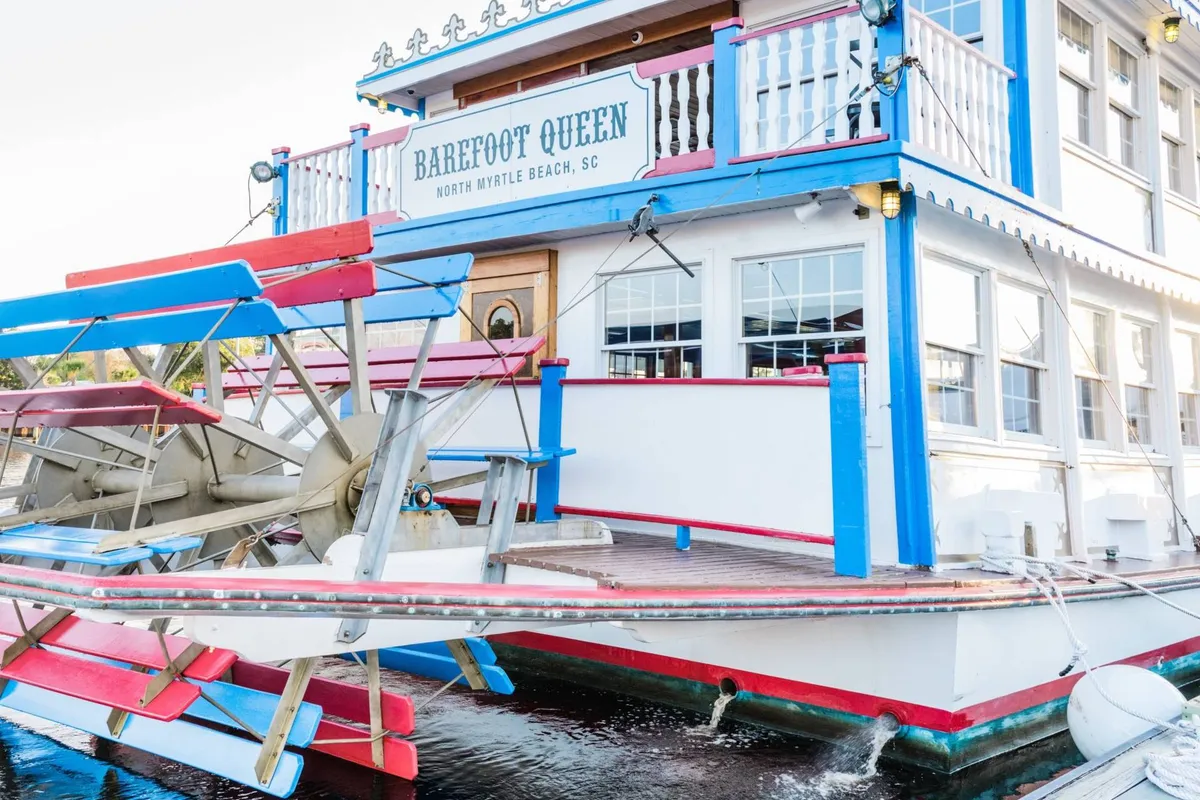 A vibrant paddle wheel vessel with a colorful exterior is docked along the coastline, featuring prominent signage. The lively scene suggests dining, sightseeing, and live music possibilities, with the coastal backdrop hinting at both relaxation and entertainment.