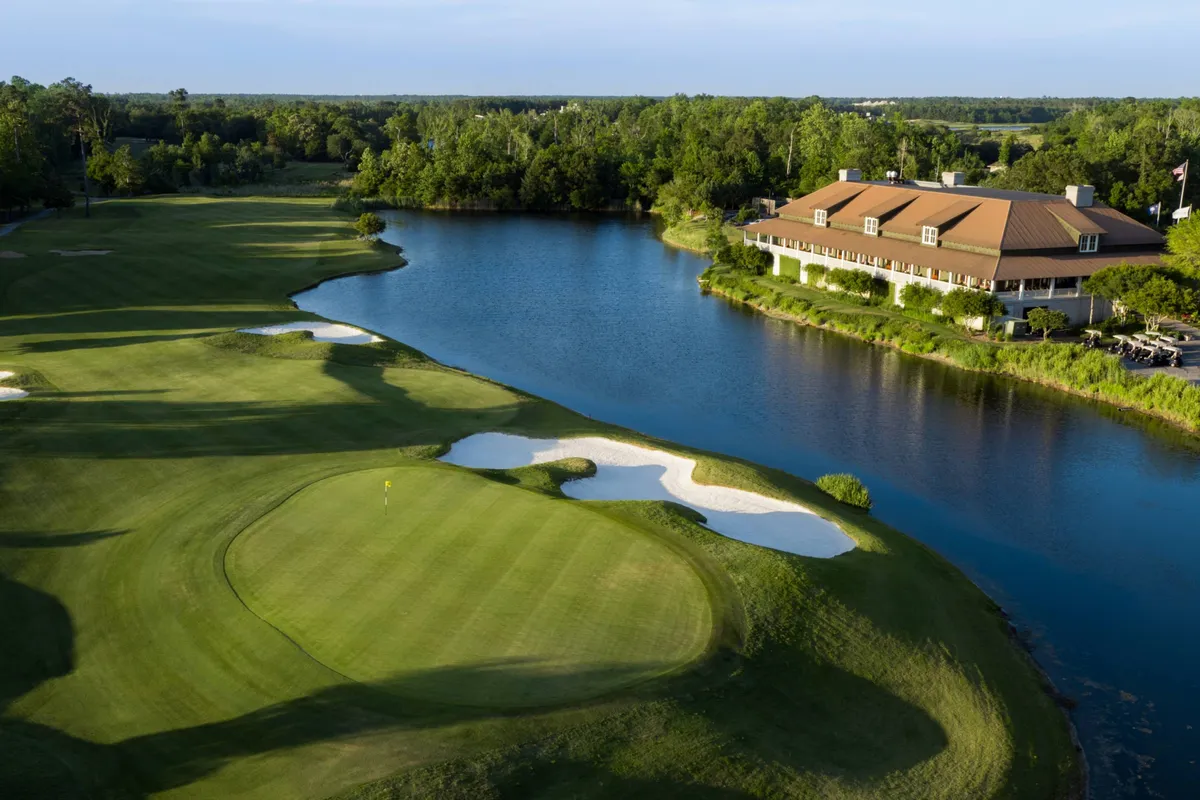 This image captures a lush golf course bordered by a serene water body, with a clubhouse in the distance and sand traps punctuating the green. The setting is open and inviting, ideal for friendly golf games, relaxing on the patio, or appreciating sunsets over the water.