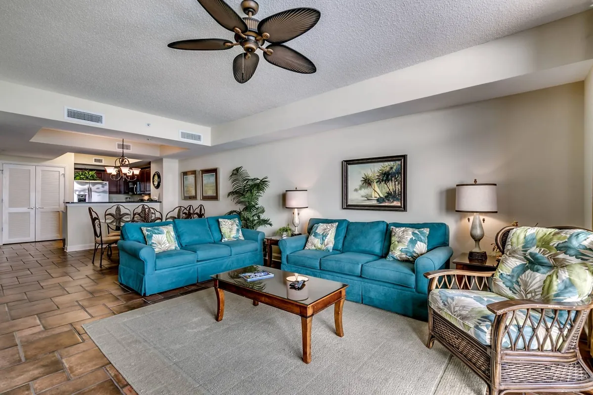 A sunlit living room featuring turquoise sofas arranged around a wooden coffee table, with tropical decor and a ceiling fan overhead. The space feels open and inviting, ideal for gatherings, relaxation, and lively conversation. Large windows hint at the ocean views beyond, reinforcing the seaside atmosphere.