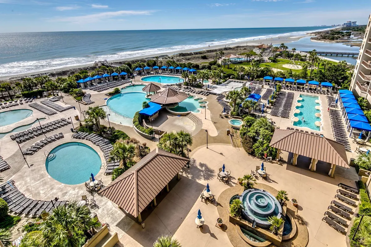 A wide exterior view of the resort's pool complex, featuring multiple swimming pools bordered by palm trees, lounging areas, easy access to the beach, and barbecue areas. Guests of all ages are seen enjoying the water and the sun, with spaces for both family gatherings and quiet relaxation. The image highlights the resort’s vibrant poolside life and scenic ocean views.