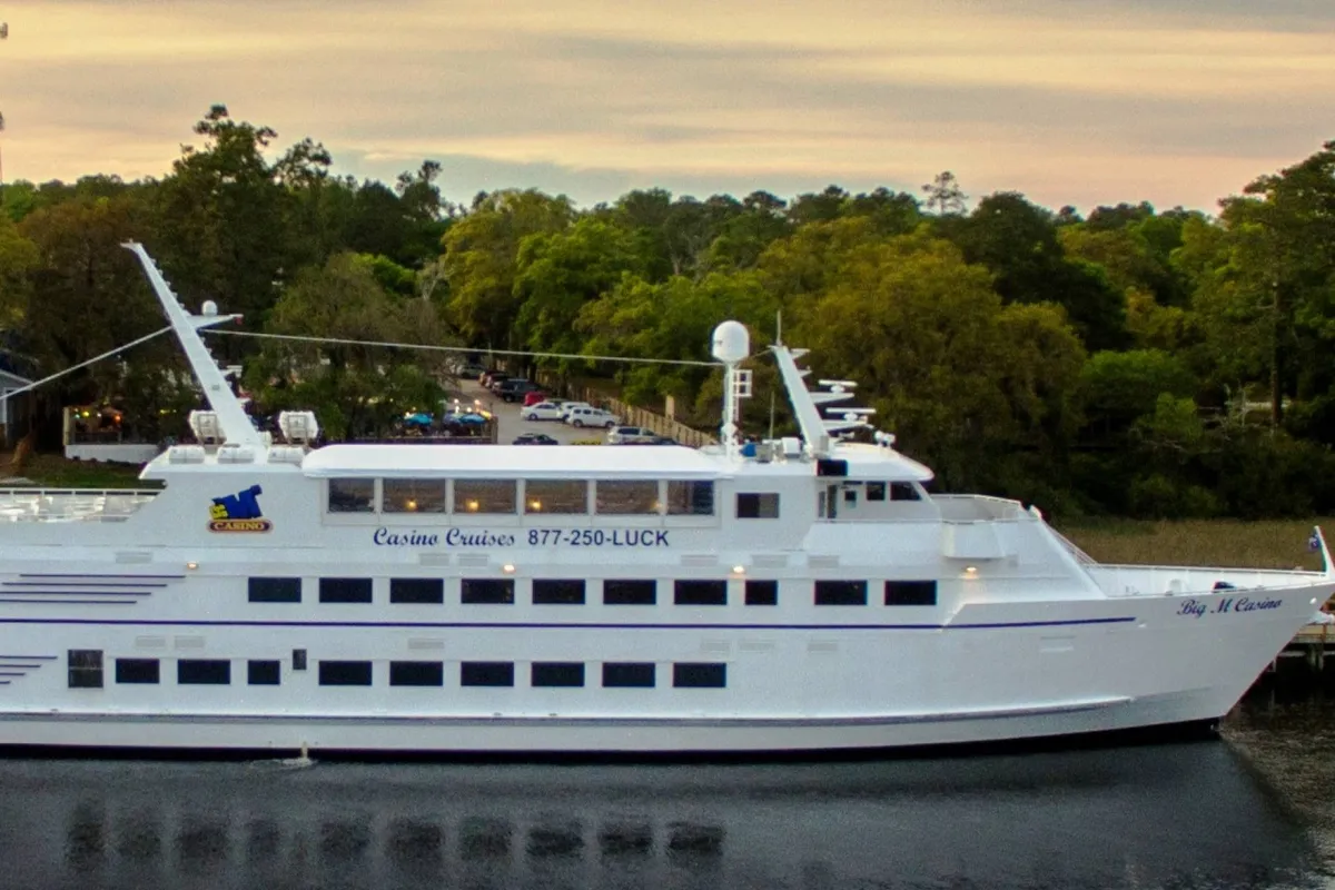 A brightly painted casino cruise ship sits among trees, its signage promising gaming and entertainment. The festive exterior hints at lively on-board experiences, including dining, gambling, and live performances, aligning with a spirited nightlife atmosphere.